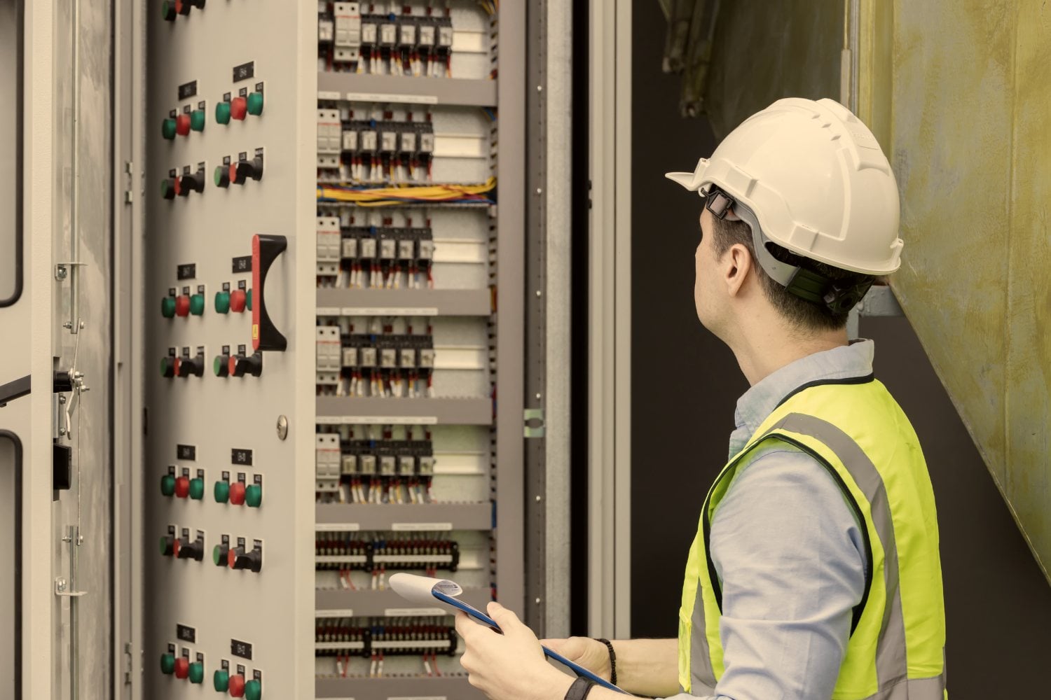 An engineer in a hard hat and hi-vis vest checking a control panel against a tablet.