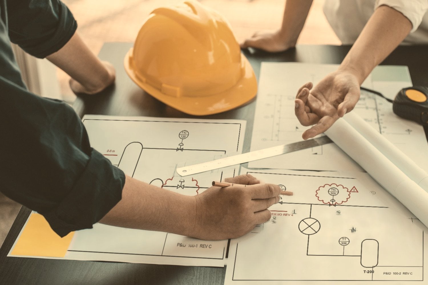 A yellow hard hat on a table covered in marked-up P&IDs, with red revision clouds, while two engineers work through them.