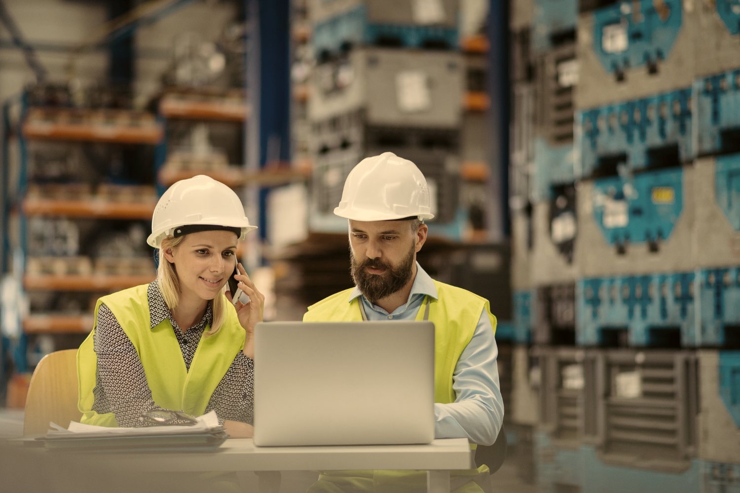 Two engineers in hi-vis vests at a laptop in a warehouse, one of them on the phone.