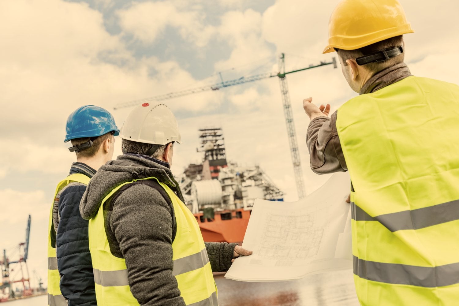 Three people in hard hats and hi-vis vests on site, one holding a drawing and another pointing out across the yard.
