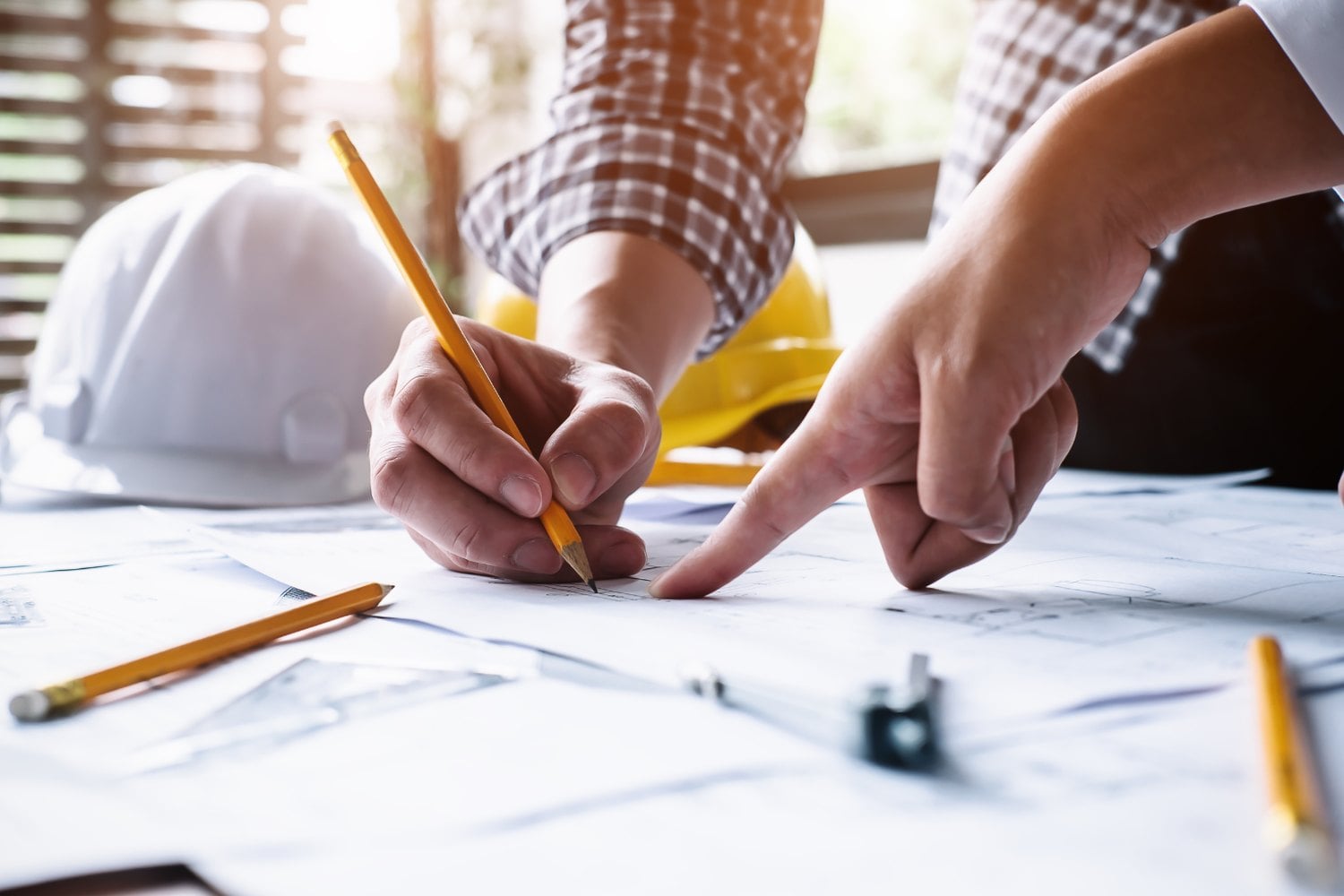 Two engineers at a table of drawings, one writing with a pencil and the other pointing, with hard hats beside them.