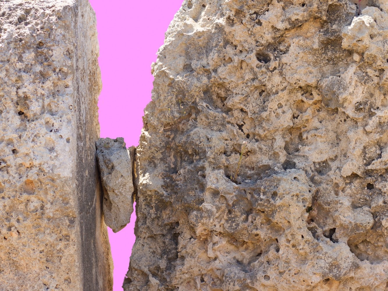 Two huge blocks of weathered stone with a small stone wedged between them, against a bright pink background.