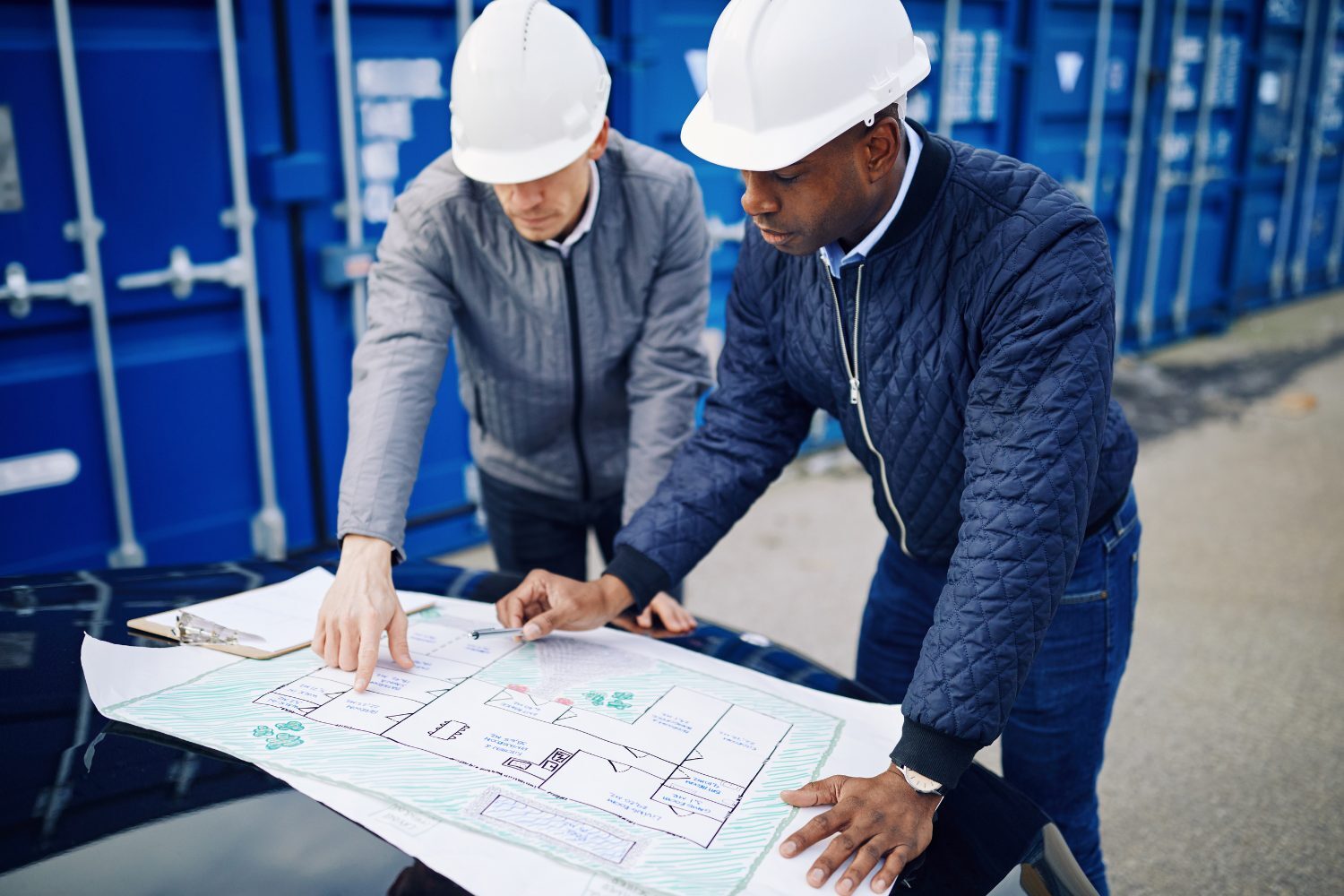 Two people in hard hats leaning over a plan drawing spread on a table, comparing it.