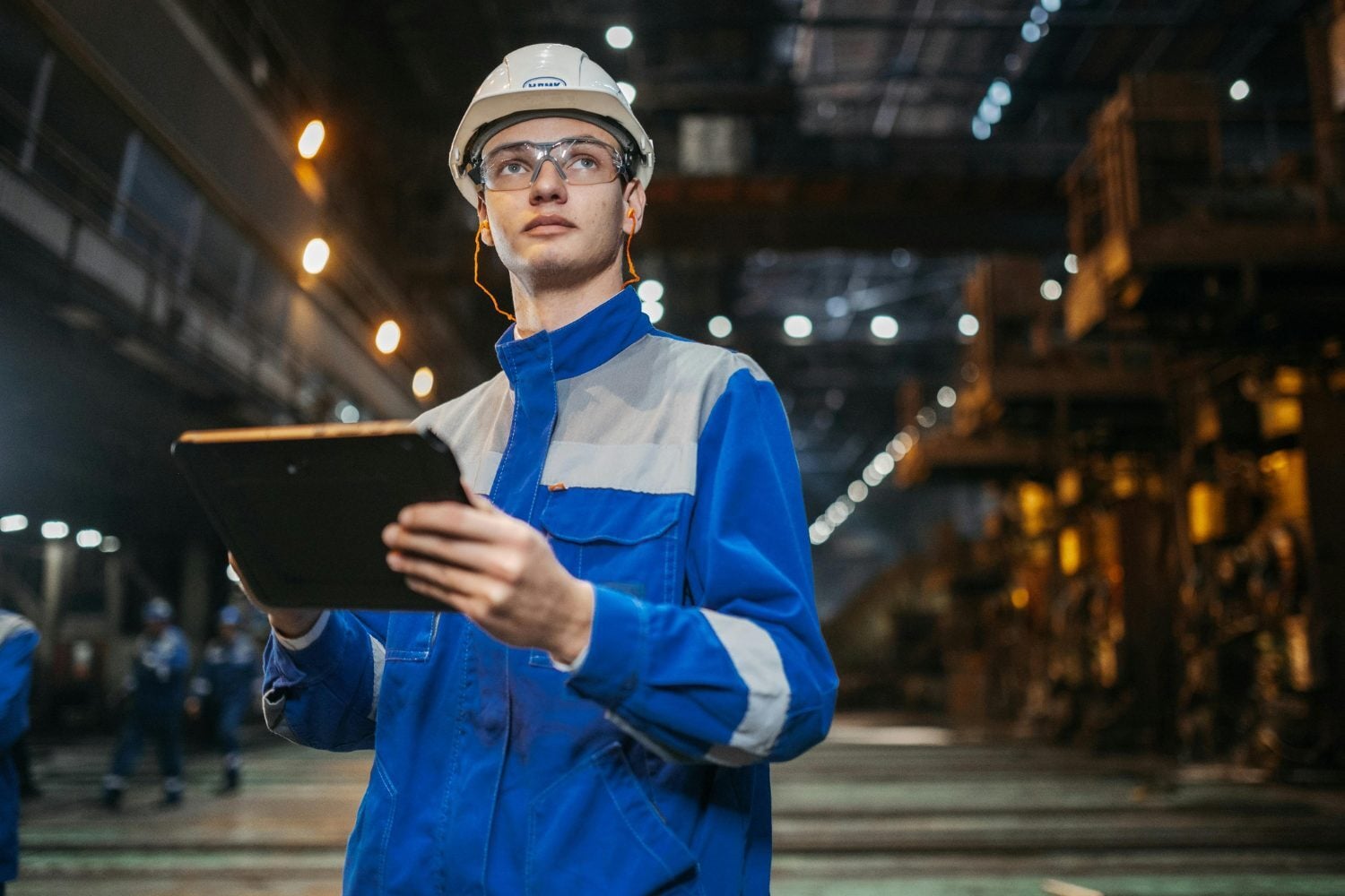 An engineer in a hard hat holding a tablet in a large industrial hall, with equipment lit up behind.